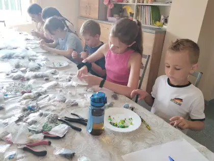 Dzieci siedzą przy dużym stole, na którym leżą w woreczkach kolorowe koraliki. Dzieci robią biżuterię z koralików.