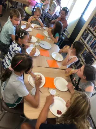 dzieci siedzą przy wspólnym stole i jedzą hot dogi.