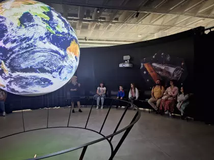W środku ciemnego pomieszczenia wisi ogromny model kuli ziemskiej. Wokoło siedzą ludzie i patrzą na planetę Ziemię.