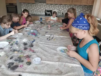 Dzieci siedzą przy dużym stole, na którym leżą kolorowe koraliki w małych woreczkach. Dzieci robią biżuterię z koralików.