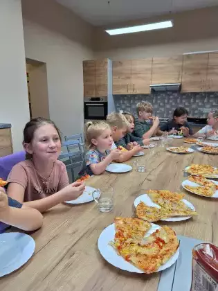 Dzieci siedzą przy wspólnym stole i jedzą pizzę.