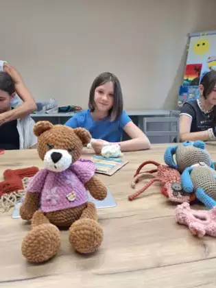 Uczestniczka warsztatów poznaje podstawy szydełkowania. Przed nią na stole siedzi misiu zrobiony na szydełku.