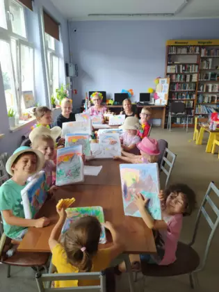 Uczestnicy siedzą przy stole i prezentują swoje namalowane obrazy.