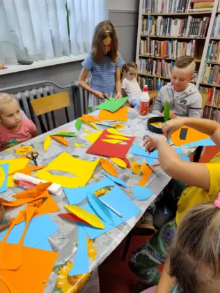 Dzieci siedzą przy stoliku i wycinają pióropusze z kolorowego papieru.