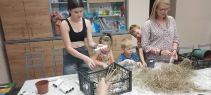 Dzieci siedzą przy stole i wypełniają doniczki sianem i szyszkami.
