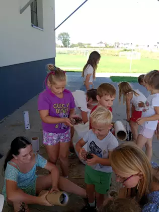 Dzieci kucają na dworze i wypełniają tekturowe rolki sianem i szyszkami.