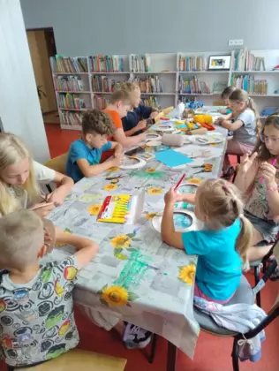 Dzieci siedzą przy stole i kolorują kredkami naszyjniki afrykańskie.