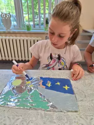 Dziewczynka maluje obrazek kolorowymi mazakami. Obrazek przedstawia góry, drzewa i wilka wyjącego do księżyca.