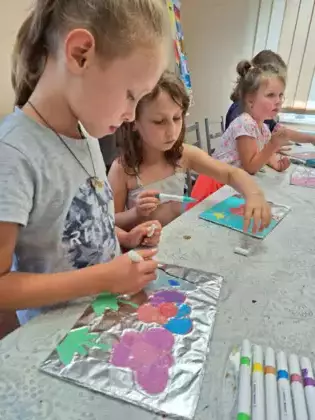 Dziewczynka maluje obrazek na folii przedstawiający kwiaty rosnące na trawie.