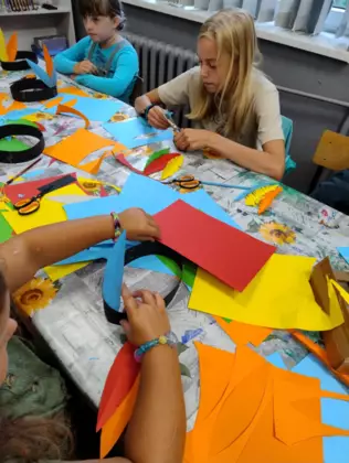 Dzieci siedzą przy stoliku i wycinają pióropusze z kolorowego papieru.