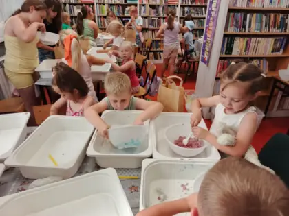 Dzieci siedzą przy stolikach i w miskach mieszają kolorowy piasek kinetyczny.
