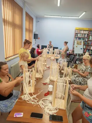 Uczestnicy warsztatów makramy siedzą przy długim stole w bibliotece i wykonują dekoracje ze sznurka na drewnianych ramach. W zajęciach biorą udział dzieci, młodzież i dorośli. Przy stołach widać przygotowane materiały, nożyczki oraz kolejne etapy powstających prac.