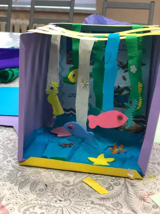 Na stole papierowe akwarium. W środku zwisają na bibule kolorowe rybki.