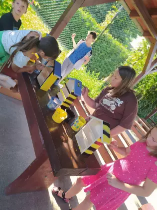 Dzieci na zajęciach stolarskich, na podwórku pod daszkiem. Dzieci w wieku szkolnym malują drewniane domki dla owadów w kolorach żółty, niebieski i czarny. Jest słoneczna pogoda.