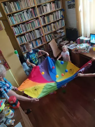 W bibliotece czwórka dzieci w wieku szkolnym trzyma kolorową chustę. W chuście są kolorowe piłeczki. Dzieci się bawią.