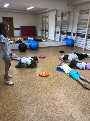 Dzieci ćwiczą na hali sportowej. Jedna ściana jest z luster. Część dzieci leży na brzuchu. Jedna dziewczynka stoi.