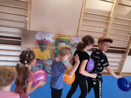 Kilka dzieci stoją jeden za drugim, trzymają kolorowe balony. Duży karton oparty o ścianę a na nim namalowana lokomotywa.