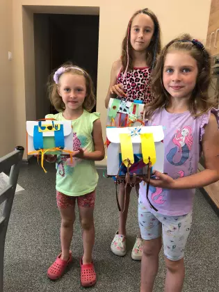 Trzy kilkuletnie dziewczynki pozują do zdjęcia, w rękach trzymają papierowe plecaki szkolne, uśmiechają się.