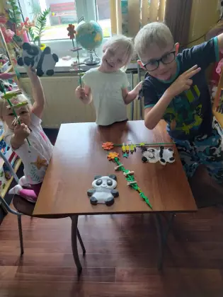 W bibliotece dzieci w wieku przedszkolnym stoją uśmiechnięte przy stoliku. Na stoliku leżą kolorowe prace plastyczne, gipsowe misie panda i papierowe kwiatki na patyku wykonane przez dzieci.