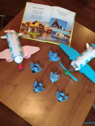 Na stole leży otwarta książka a w niej zdjęcia z Australii, opery w Sydney. Obok książki leżą zrobione samoloty z plastikowych butelek oraz gipsowe pszczoły pomalowane na niebiesko przez dzieci.