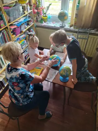 Dzieci w wieku przedszkolnym w bibliotece przy stole patrzą na globus. Trzymają ręce na globusie. 