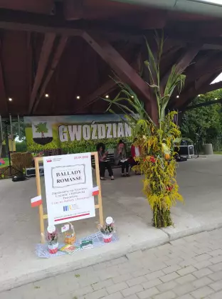 Scena pod wiatą. Z przodu duży biały plakat Narodowe Czytanie 2022. Z Tyłu na ściance napis Gwoździany.