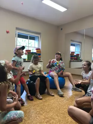 Grupa dzieci na hali sportowej. Część siedzi na piłkach dmuchanych. Po prawej stronie jest lustro na ścianie.