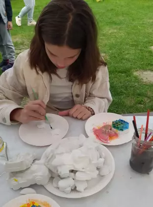 Dziewczynka w ciemnych włosach siedzi przy stole maluje białe gipsowe figurki.