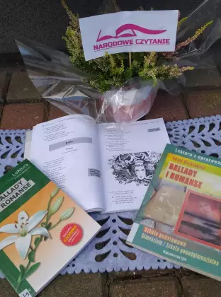 Na deskach niebieski obrus i trzy książki Adama Mickiewicza. Jedna książka jest otwarta. Z tyłu wrzosy w doniczce owinięte przezroczystą folią.