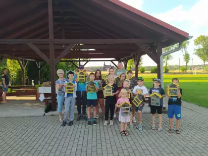 Przed dwuspadową wiatą stoi grupa dzieci szkolnych wraz z panią opiekunką oraz instruktorem stolarstwa. Dzieci trzymają drewniane kolorowe domki dla owadów. Domki są w kolorach: żółty, niebieski i czarny. Jest słonecznie, 