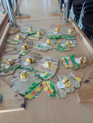 Na stole leżą prace plastyczne. Na kawałkach z kartonu w owalnym kształcie żółte pszczółki z przyklejonymi białymi skrzydełkami.