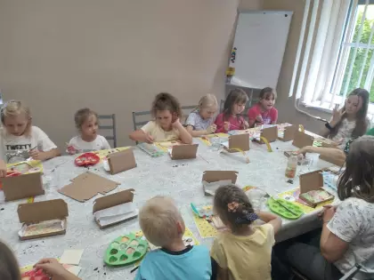 Dzieci siedzą przy stole i dekorują manufakturę czekolady. Obok leżą kartonowe pudełka do pakowania.