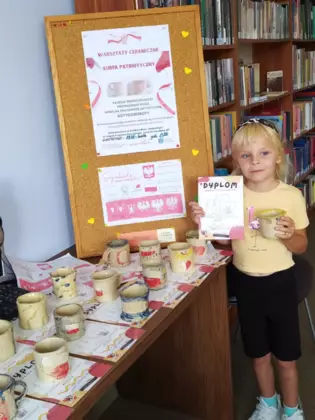 Dziewczynka stoi na tle gazetki i trzyma w ręce kubek i dyplom.