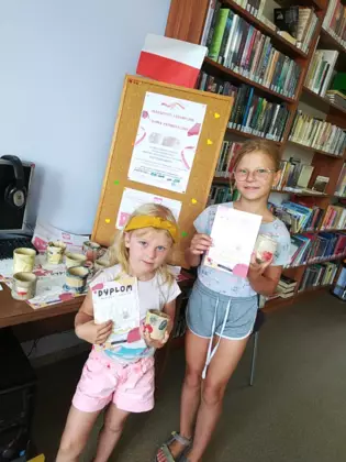 Dziewczynki stoją w bibliotece trzymając w ręce dyplom i kubek.