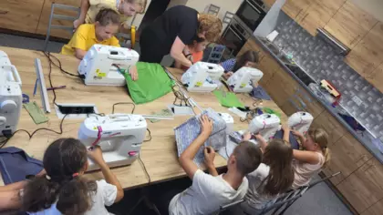 Dzieci siedzą przy stole przy maszynach i szyją workoplecaki.