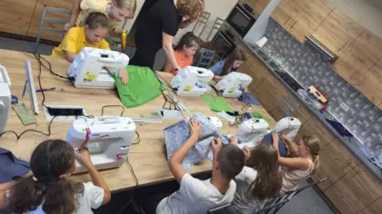 Dzieci siedzą przy stole przy maszynach i szyją workoplecaki.