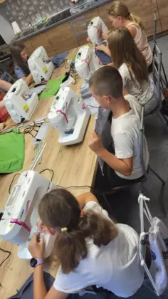 Dzieci siedzą przy stole przy maszynach i szyją workoplecaki.