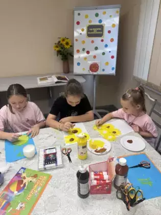 Trzy dziewczynki siedzą przy stole i malują farbami słoneczniki.
