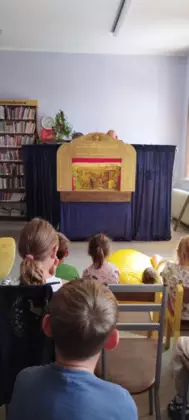 grupa dzieci siedzi i ogląda aktorów z marionetkami na scenie w bibliotece.