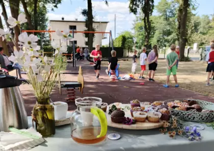 Stół zastawiony babeczkami i kawą w tle widać dzieci bawiące się na placu zabaw.
