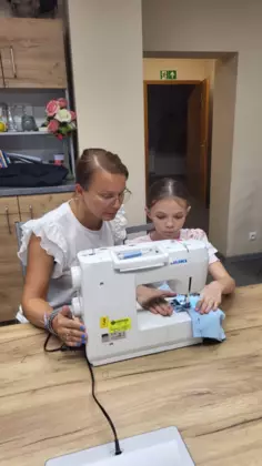 Dziewczynka wraz z instruktorką siedzi przy maszynie do szycia.
