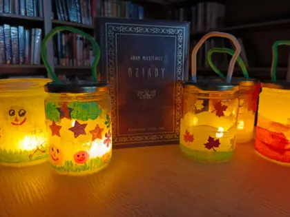 Cztery świecące lampiony stoją na stoliku i pośrodku książka Dziady Adama Mickiewicza