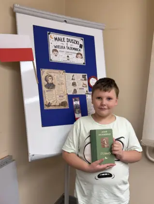 Chłopiec uczestniczący w zajęciach w Bibliotece w Pawonkowie pozuje z książką „Dziady” Adama Mickiewicza. Trzyma książkę w dłoniach i prezentuje ją przed biblioteczną gazetką poświęconą autorowi i jego utworowi.