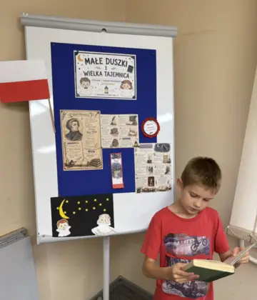 Chłopiec podczas zajęć w Bibliotece w Pawonkowie czyta fragment utworu „Dziady” Adama Mickiewicza. Stoi przed biblioteczną gazetką poświęconą Adamowi Mickiewiczowi i „Dziadom”. W rękach trzyma książkę i odczytuje fragment tekstu przed grupą uczestników.