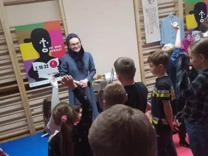 Siostra Misjonarka a wokół niej grupka kilkunastoletnich dzieci. Z tyłu na ścianie drewniane drabinki, na których powieszone są plakat i kolorowe ubrania z Tajwanu. 