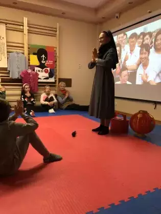 Sala sortowa, na środku stoi Siostra Misjonarka ze złożonymi dłońmi. Za nią jest duży ekran na którym wyświetla się prezentacja o Tajwanie. Dzieci siedzą na podłodze, patrzą na Siostrę.