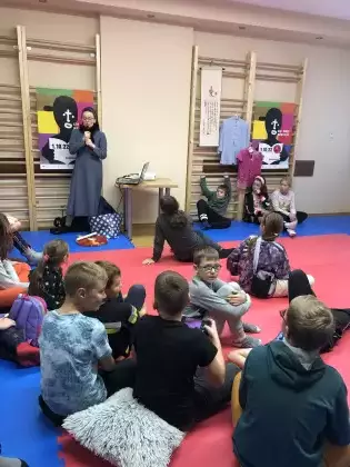 Dzieci siedzą w sali sportowej na podłodze. Patrzą w stronę Siostry Misjonarki. Na ścianie drewniane drabinki, na drabinkach powieszone duże kolorowe plakaty i ubrania Tajwańskie.