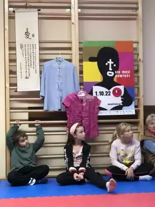 troje dzieci siedzi na sali gimnastycznej pod drewnianymi drabinkami. Na drabinkach są powieszone Tajwańskie ubrania i plakat Noc Bibliotek.