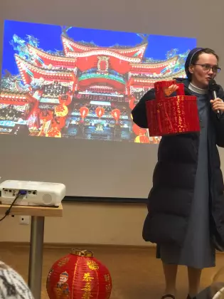 Siostra zakonna stoi przy ekranie, w rękach trzyma mikrofon i czerwony kuferek . Na ekranie prezentacja z chin. 