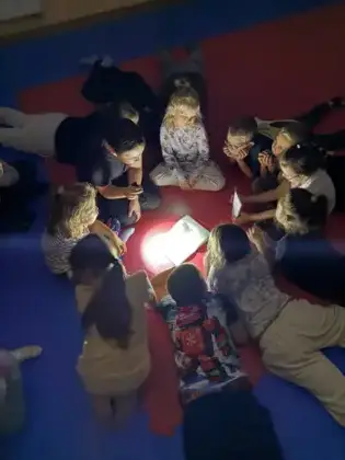 Dzieci leżą na materacach i słuchają z uwagą czytanej książki przy świetle latarki, w tajemniczej i magicznej atmosferze Nocy Bibliotek.